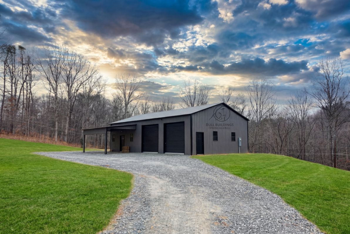 Dark gray metal building with three garage doors and a covered entry on a gravel driveway in a rural North Carolina setting.
